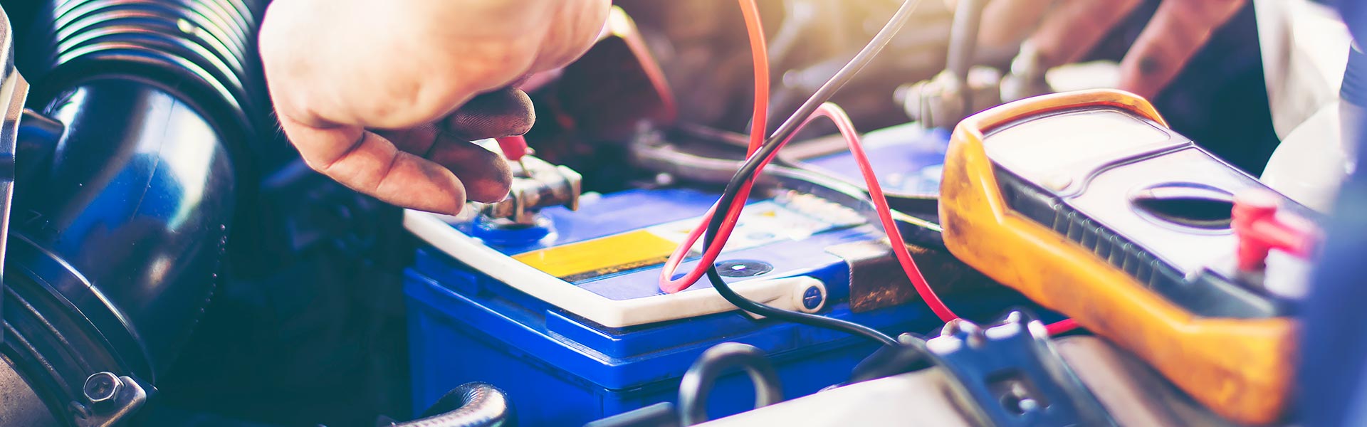 A technician using a multimeter test lead on a car battery terminal under the hood.